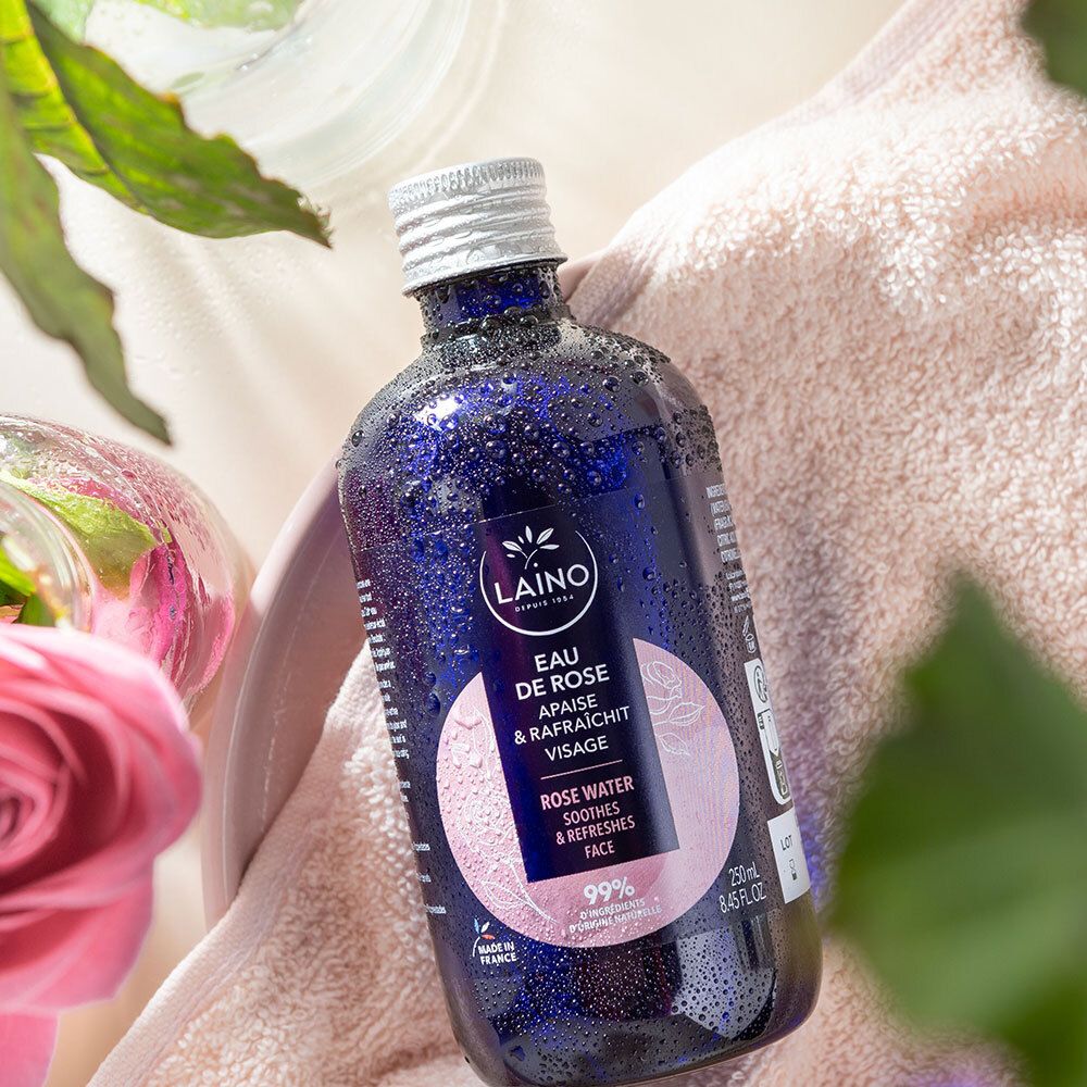 Flacon bleu avec bouchon argenté sur une serviette. Inscription: LAINO, Eau de Rose, apaise & rafraîchit le visage. Roses et verre.
