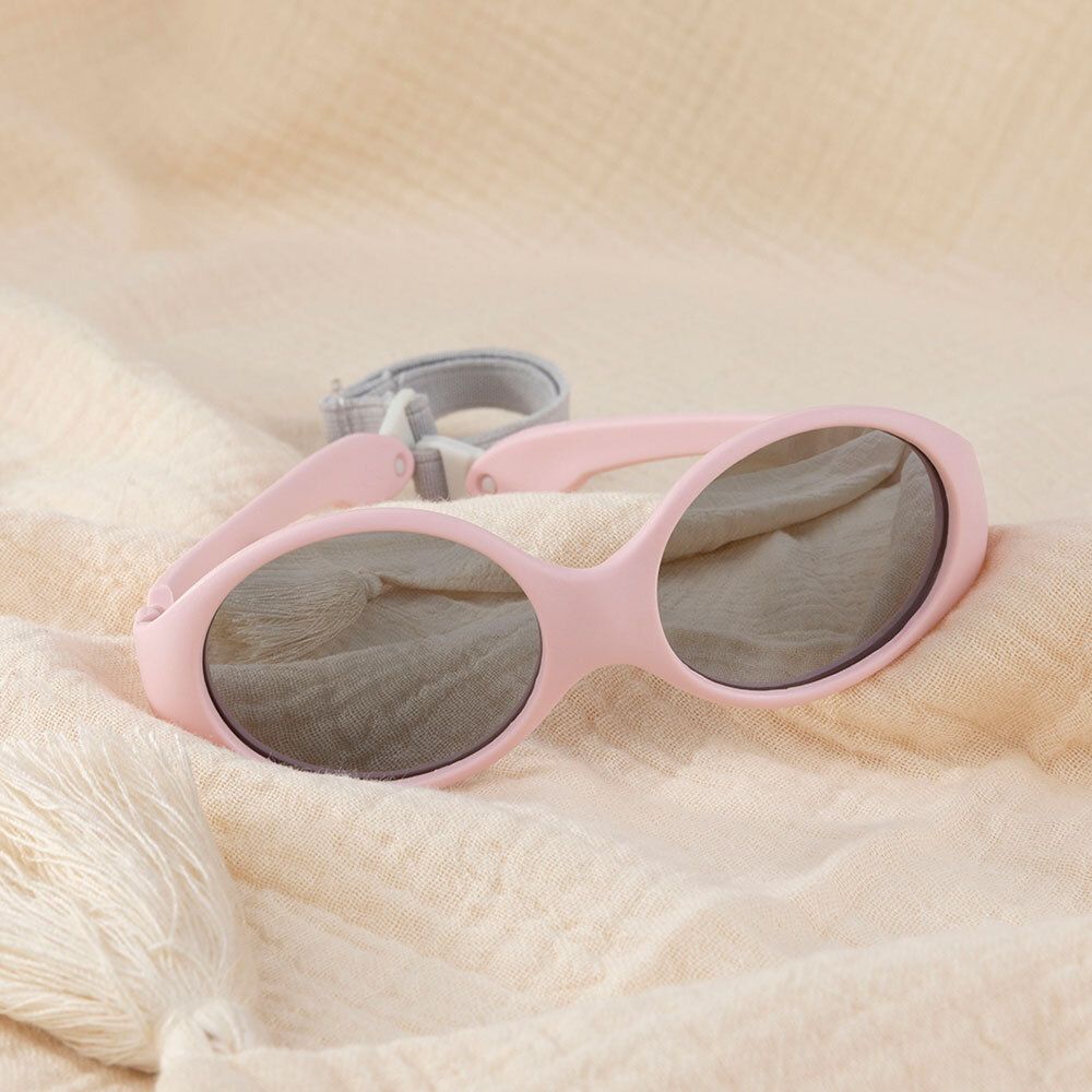 Lunettes de soleil bébé roses avec verres gris et bandeau. Sur tissu beige.