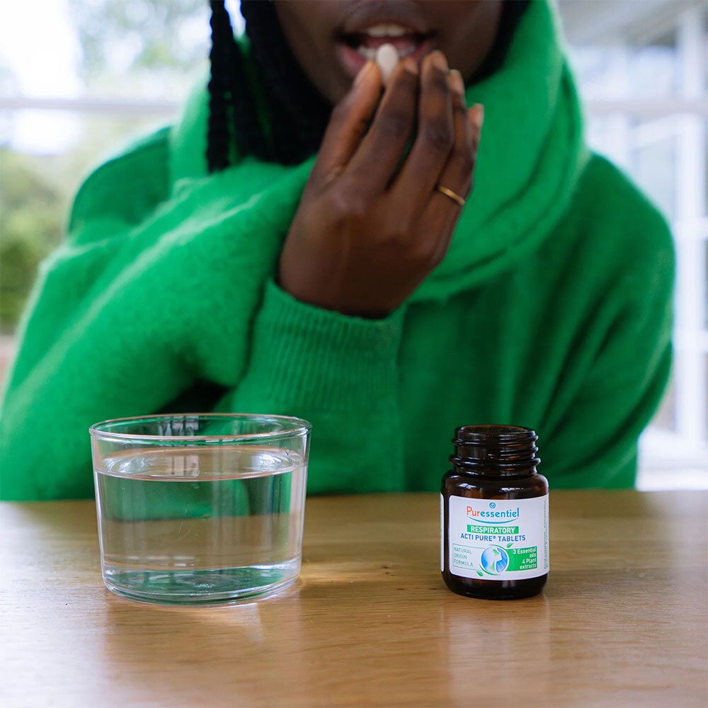 Personne prenant un comprimé. Sur la table : verre d'eau et flacon PURESSENTIEL Respiratoire Acti Pure® Comprimés.
