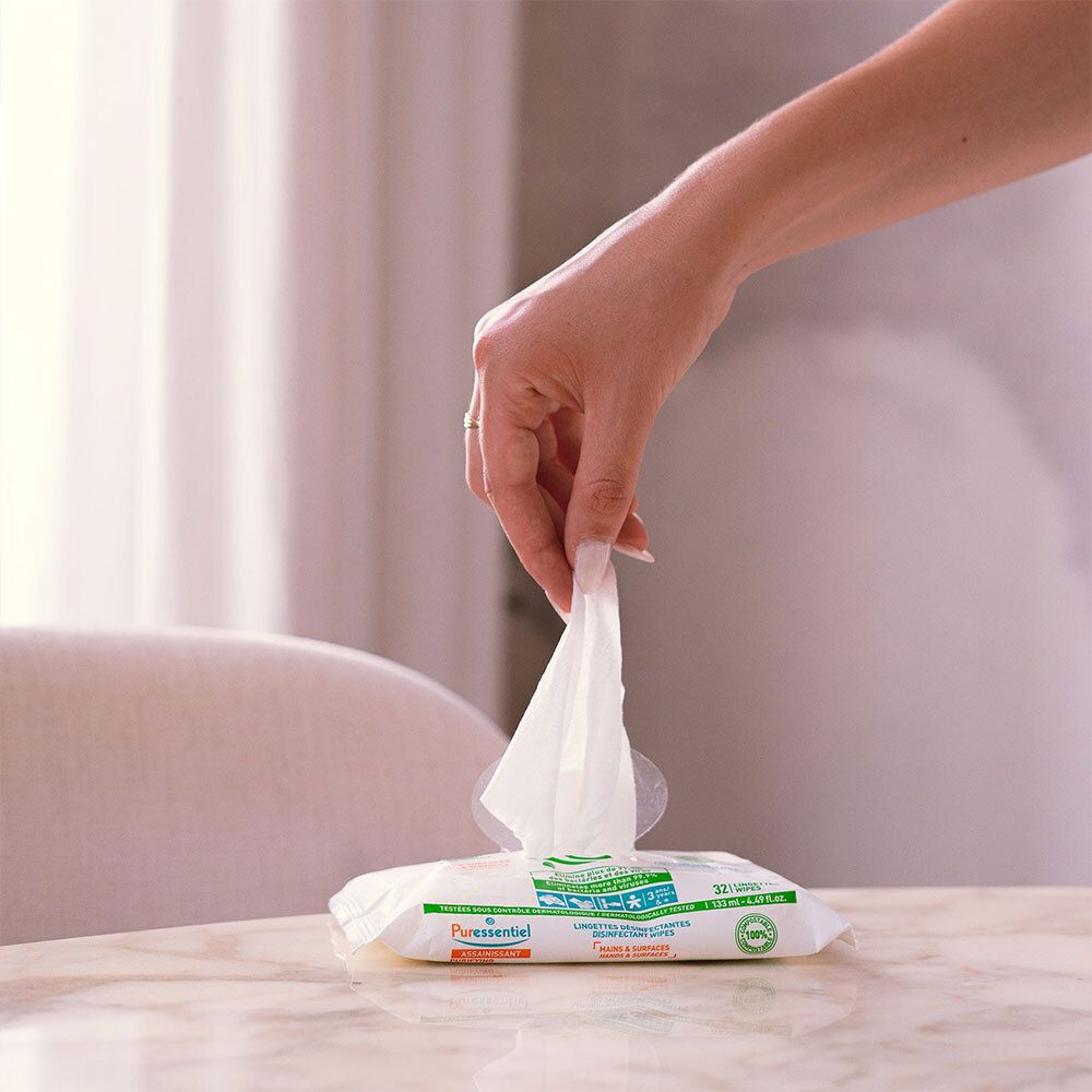 Main saisissant un paquet de lingettes désinfectantes Puressentiel. Emballage blanc, lingettes extraites. Sur table en marbre.