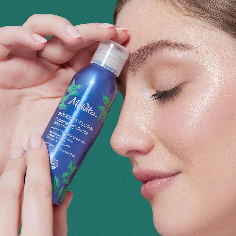 Flacon de poudre exfoliante Melvita dans un nuage de poudre blanche. Flacon bleu à feuilles vertes.