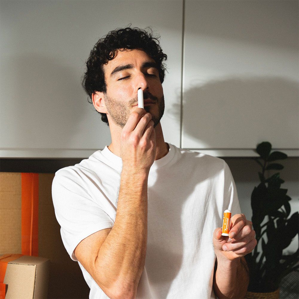 Un homme tient un inhalateur blanc et orange devant son nez. Il porte un t-shirt blanc.