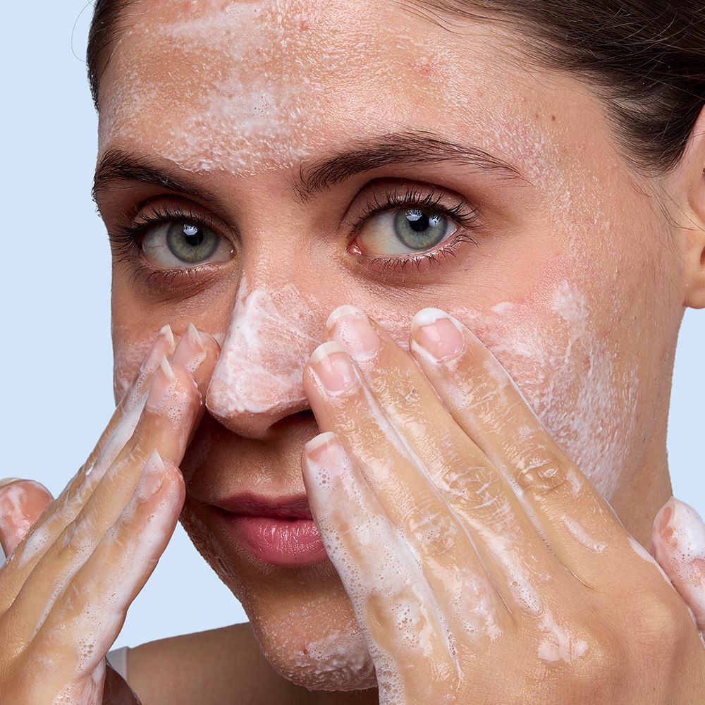 Visage couvert de mousse blanche. Mains frottant le visage. Pas de nom de produit visible.