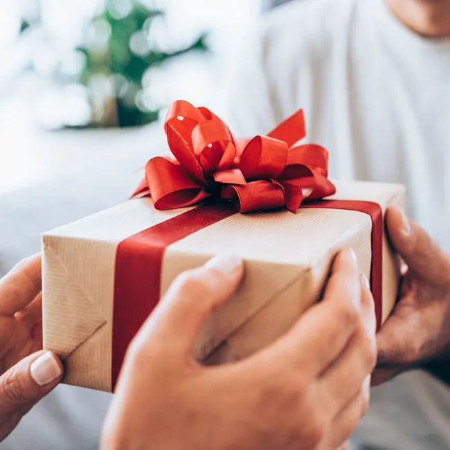 Un cadeau emballé dans du papier brun avec un ruban et un nœud rouges. Des mains tiennent le cadeau.