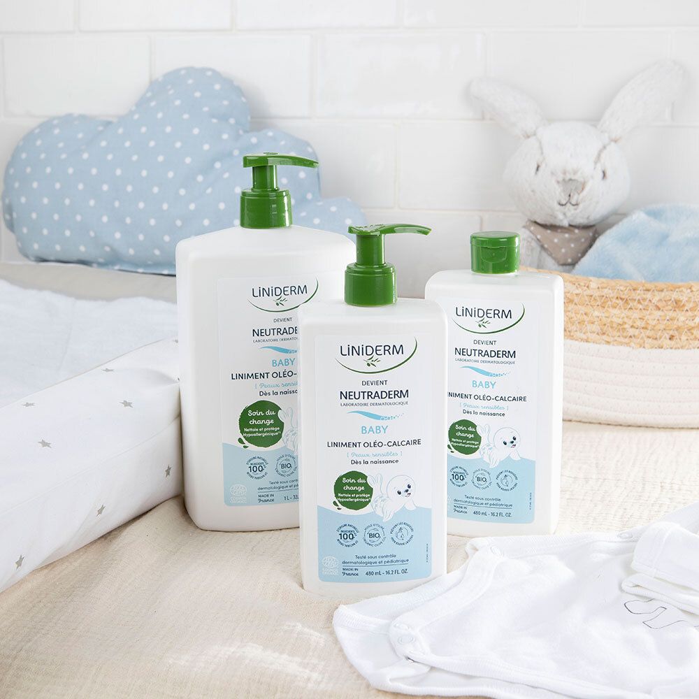 Trois flacons de liniment bébé avec bouchons verts. Sur une table, à côté de jouets et de vêtements.