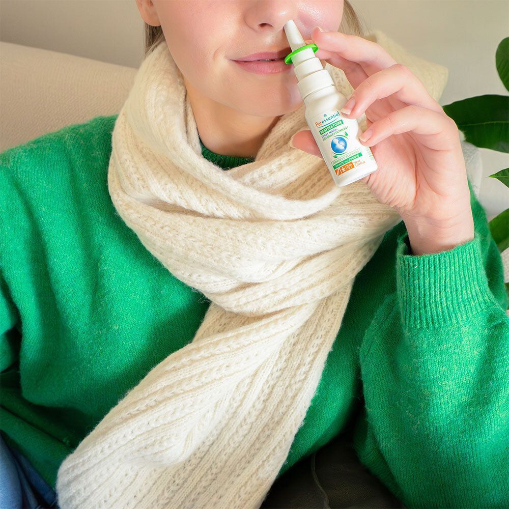 Personne tenant un spray nasal devant le nez. Spray blanc avec applicateur vert. La personne porte un pull vert et une écharpe.