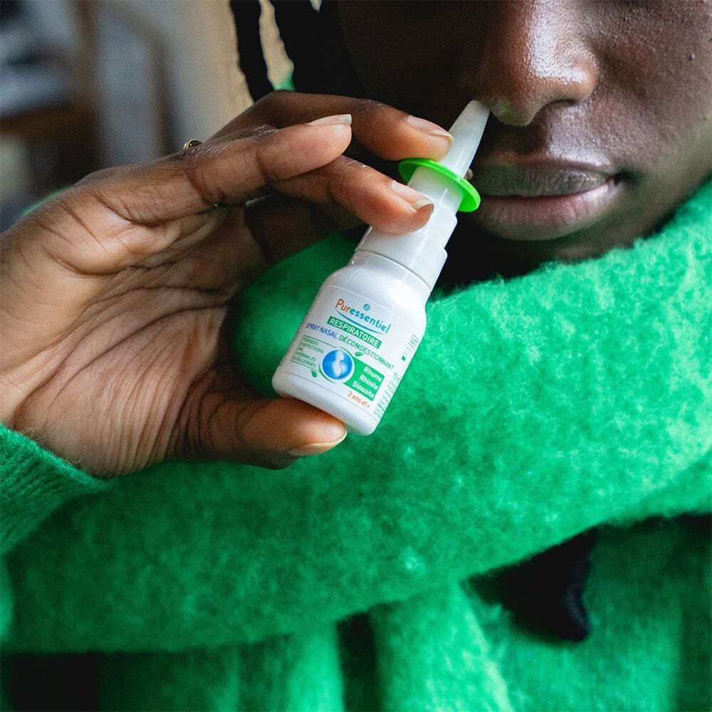 Personne tenant un spray nasal devant le nez. Spray blanc avec applicateur vert. La personne porte un pull vert.