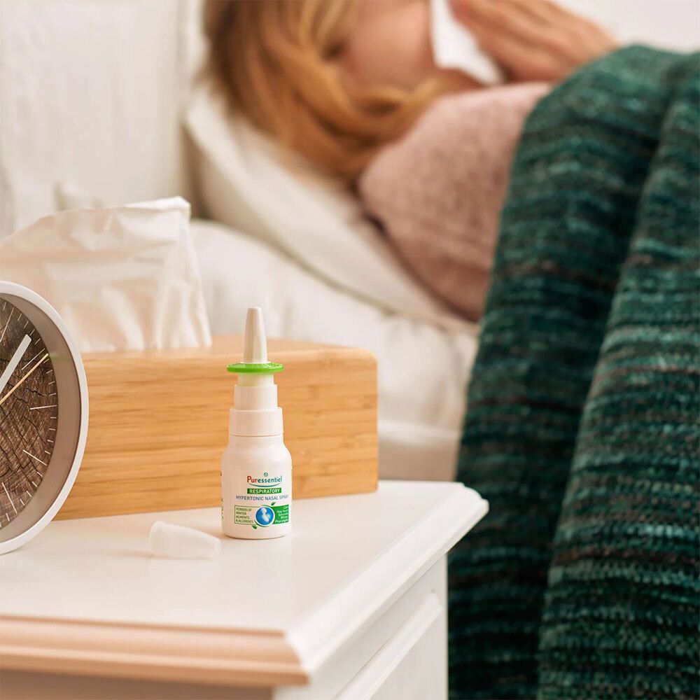 Spray nasal blanc avec applicateur vert sur table de chevet. Personne au lit en arrière-plan. Mouchoirs et horloge.