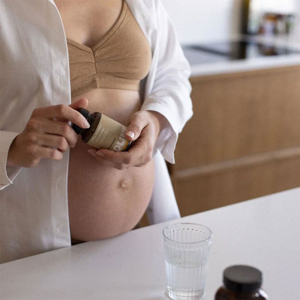 Femme enceinte tenant une bouteille marron. Sur la bouteille, il est écrit Jolly Mama Choline Mama. Un verre d'eau est à côté.