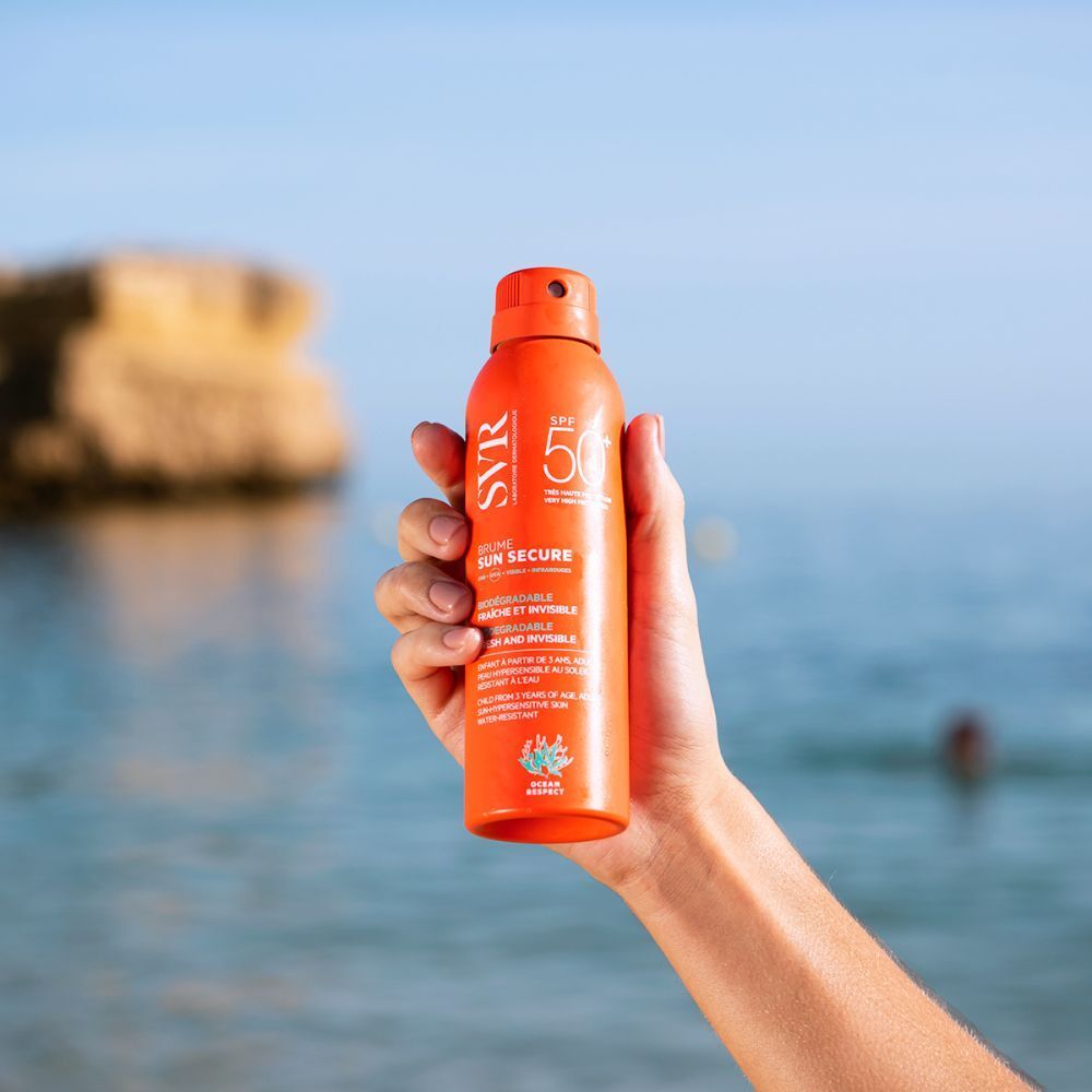 Main tenant un spray orange sur la plage. Inscription: SVR, SPF 50, Sun Secure Brume. Mer en arrière-plan.