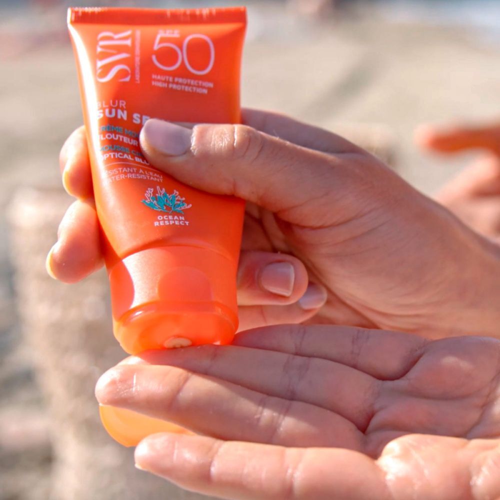 Mains tenant un tube de crème SVR Sun Secure SPF50+. Tube orange avec texte blanc. Texte: Blur, Sun Secure, Creme Mousse, Flouteur Optique, Optical Blur.