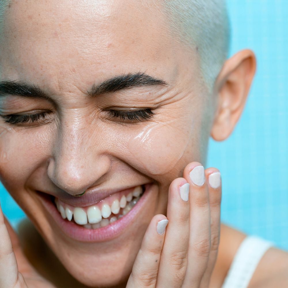 Gros plan d'une personne appliquant le produit sur son visage. Nom et logo du produit non visibles. La personne sourit.