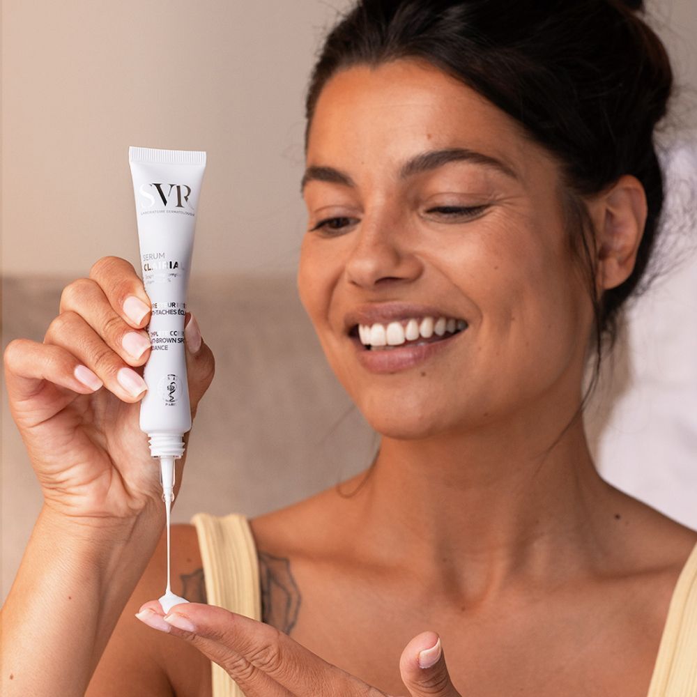 Femme appliquant du sérum SVR Clairial sur sa main. Expression souriante. Haut jaune.