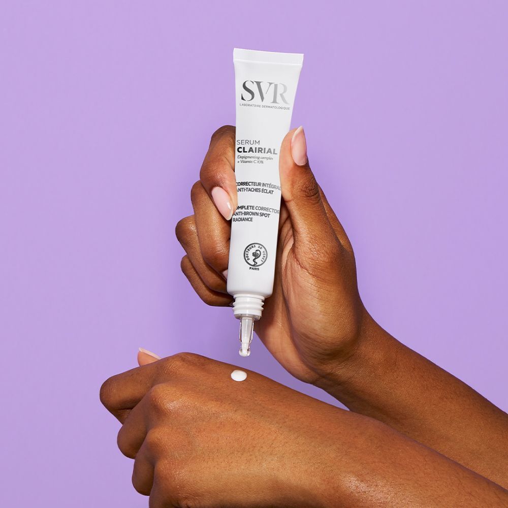Mains avec sérum SVR Clairial. Le tube est appliqué sur la main. Fond violet. Texte: Clairial Serum, Corrector intégral anti-taches éclat.