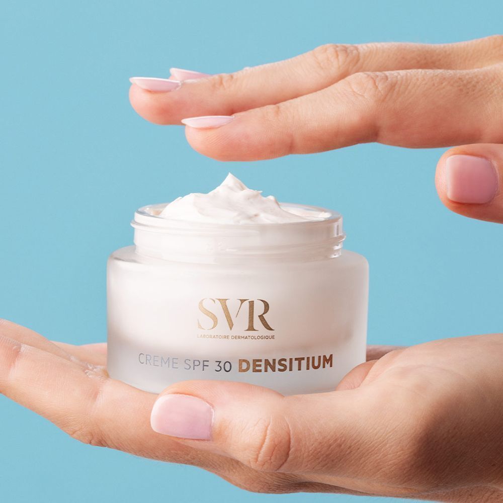 Pot de crème SVR SPF 30 DENSITIUM dans une main. Crème blanche prélevée. Fond bleu.