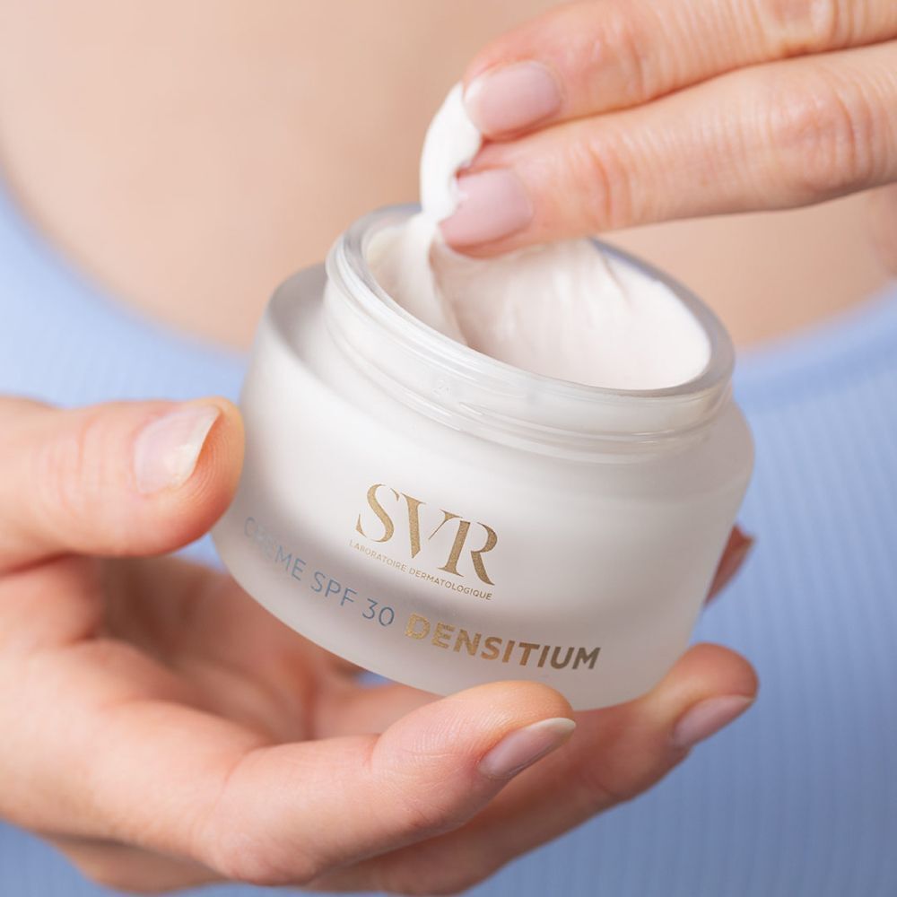 Pot de crème SVR SPF 30 DENSITIUM. Crème blanche prélevée avec un coton. Fond bleu.
