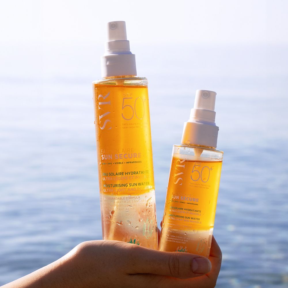 Main tenant deux flacons de SVR Sun Secure Eau Solaire SPF50+. Liquide jaune, vaporisateur blanc. Texte: SPF50+, Eau Solaire Hydratante. Mer en arrière-plan.