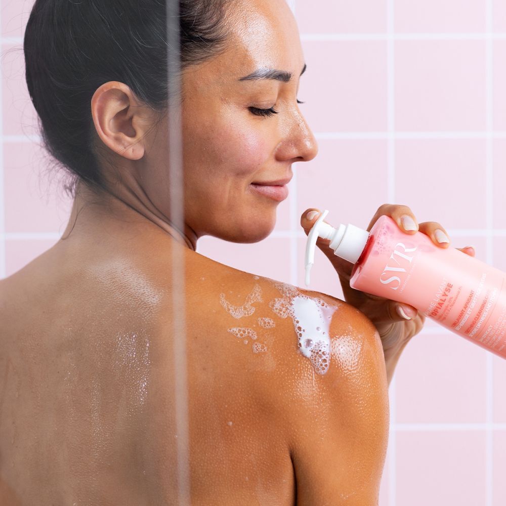 Femme sous la douche, vue de dos. Elle tient un flacon rose avec pompe blanche. Mousse sur l'épaule.