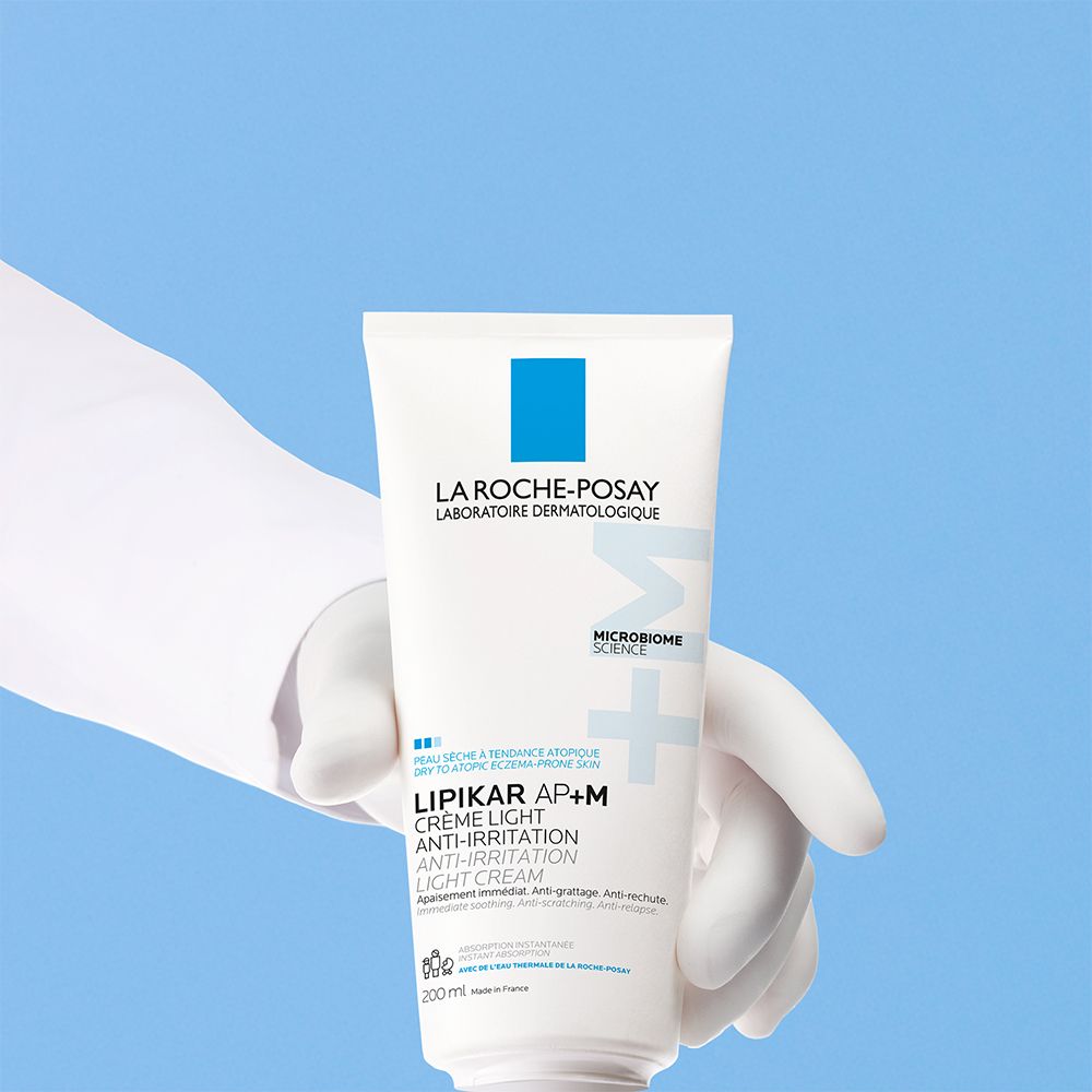 Tube blanc dans une main gantée. Texte: LA ROCHE-POSAY, LIPIKAR AP+M Crème Light Anti-Irritation. Fond bleu.