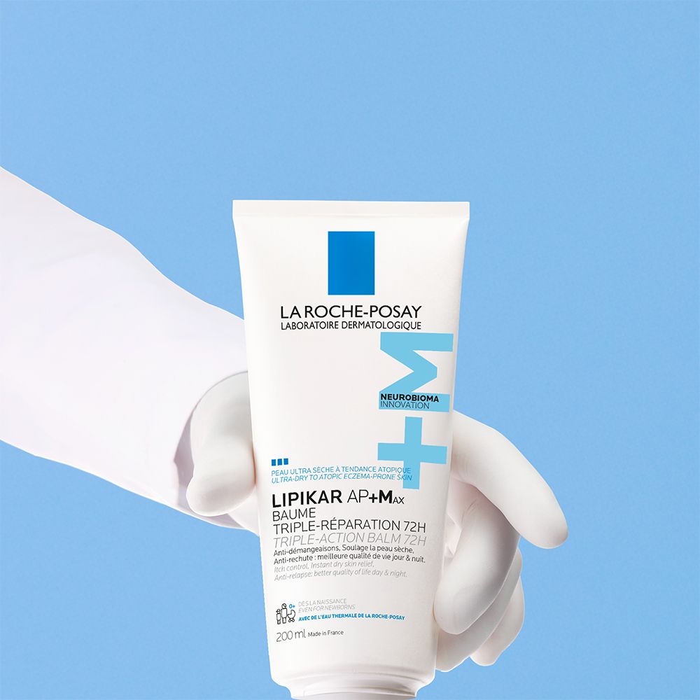 Tube blanc avec logo bleu et texte, tenu par une main gantée de blanc. Nom du produit : Lipikar Baume AP+M.