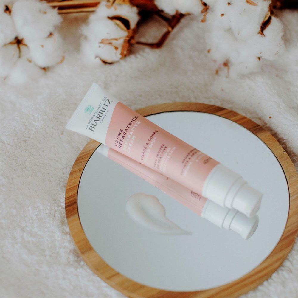 Tube de crème sur l'épaule. Design rose et blanc. Texte: Crème Réparatrice, Visage & Corps. Inscription: Peau irritée, abîmée, réactive.