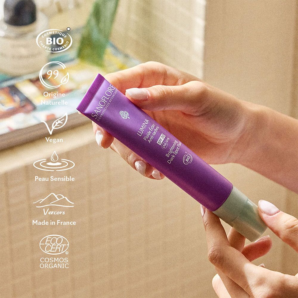 Mains tenant un tube. Texte: Lumina, SPF 30, Brightening Dark Spot Fluid. Bio, Vegan.