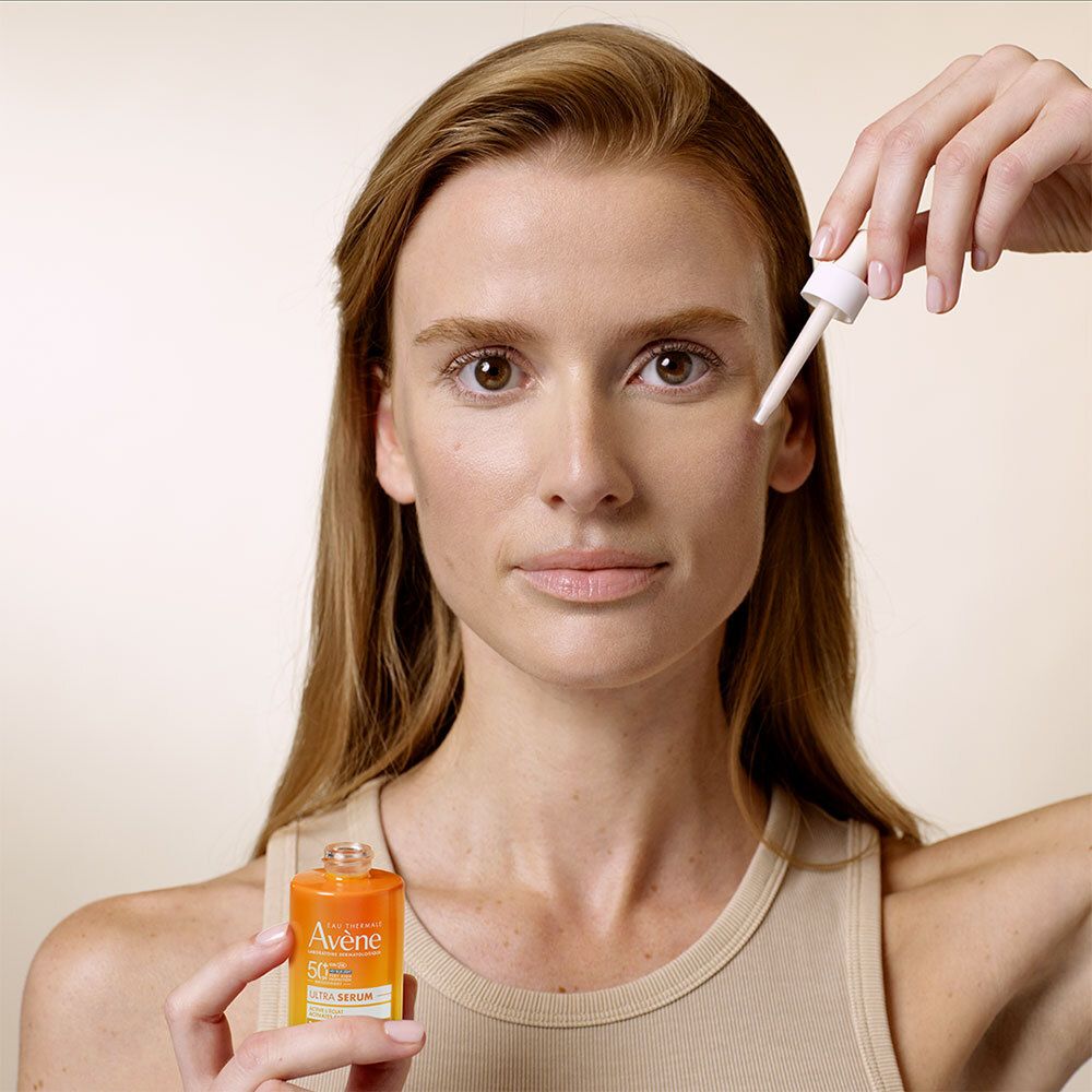 Femme tenant un flacon orange avec compte-gouttes blanc et un compte-gouttes. Inscriptions : Eau Thermale Avène, SPF 50+, Ultra Serum.