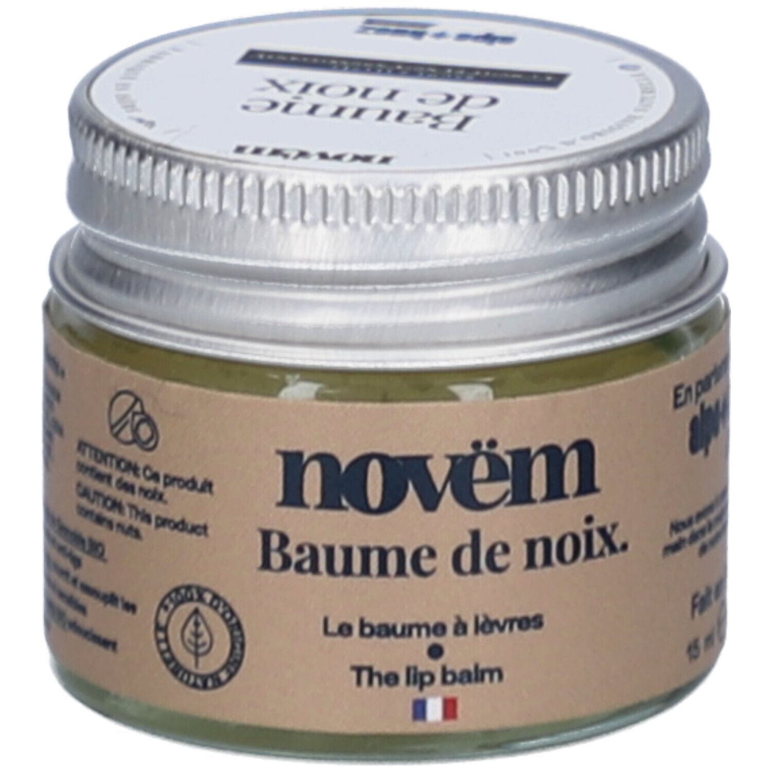 Pot en verre avec couvercle argenté. Étiquette avec nom du produit: novëm Baume de noix. Le Baume à lèvres. The lip balm. Contient des noix.