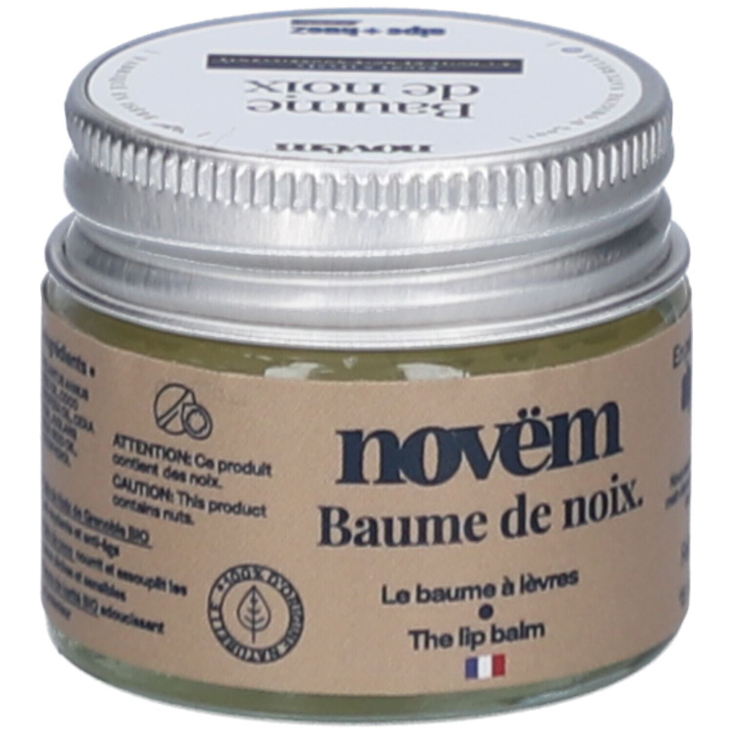 Pot en verre avec couvercle argenté. Étiquette avec nom du produit: novëm Baume de noix. Le Baume à lèvres. The lip balm. Contient des noix. Drapeau français.