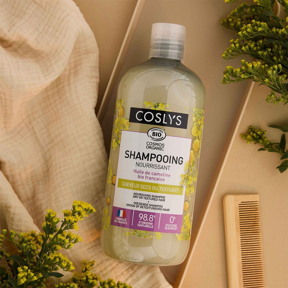 Flacon de shampooing Coslys dans une boîte. Étiquette avec informations produit. Peigne et plantes en arrière-plan.