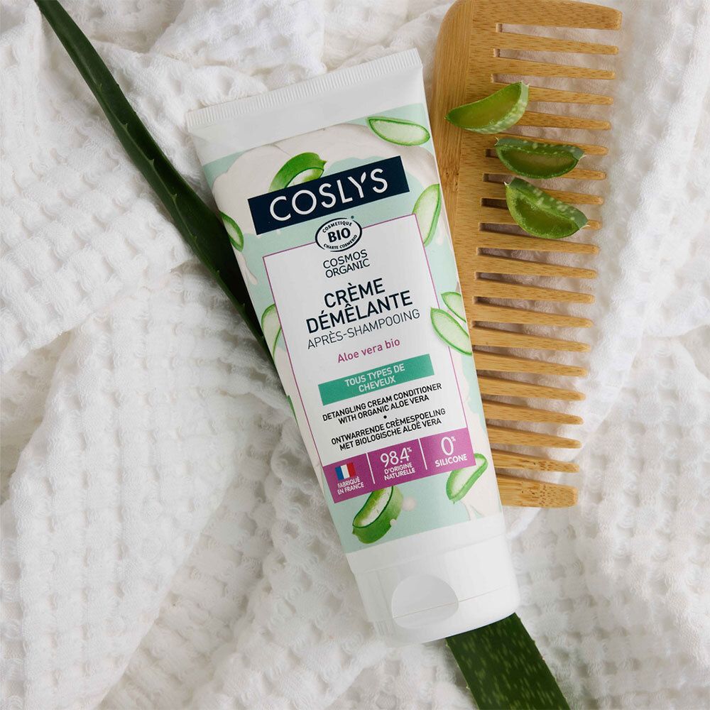 Tube de crème avec nom du produit et aloe vera, à côté d'un peigne en bois et d'une feuille d'aloe vera sur tissu blanc. Texte : Crème démêlante.