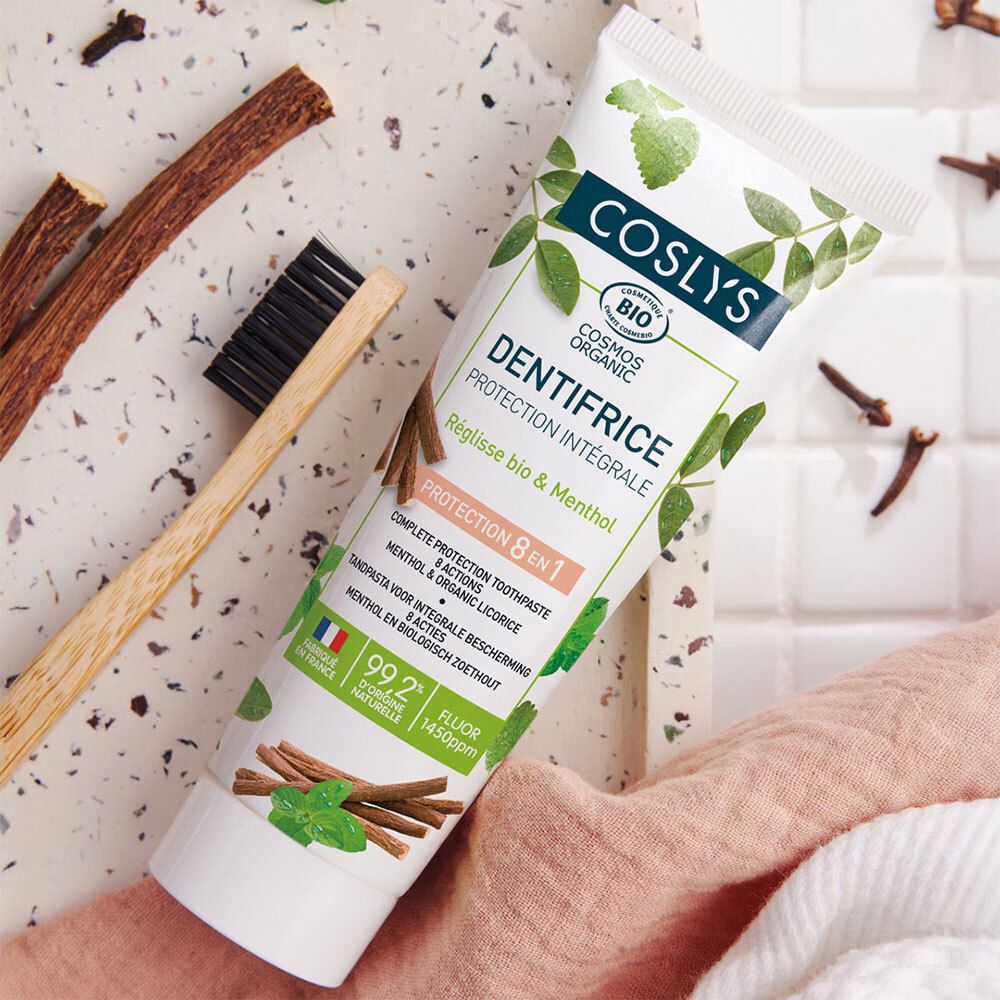 Tube de dentifrice Coslys Dentifrice Protection Intégrale, à côté d'une brosse à dents. Blanc, vert et brun. Texte : 'Protection 8 en 1'.