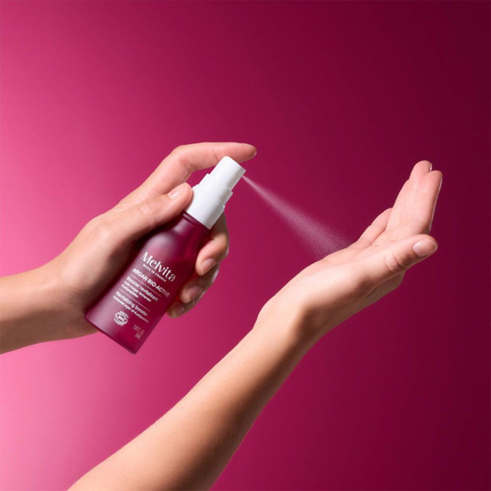 Mains vaporisant du liquide d'un flacon rouge. Inscription: Melvita Argan Bio-Active.
