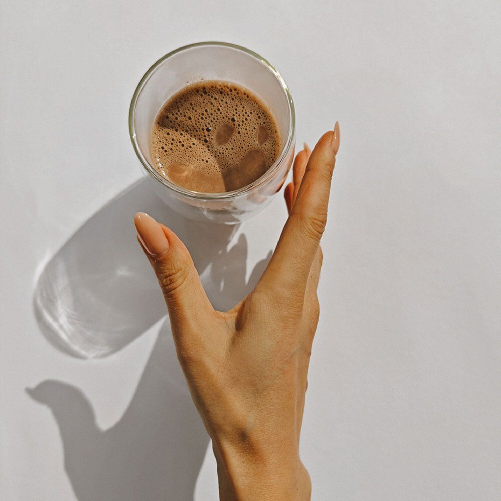 Verre contenant une boisson brune, tenu par une main avec de longs ongles clairs. Sur fond blanc.