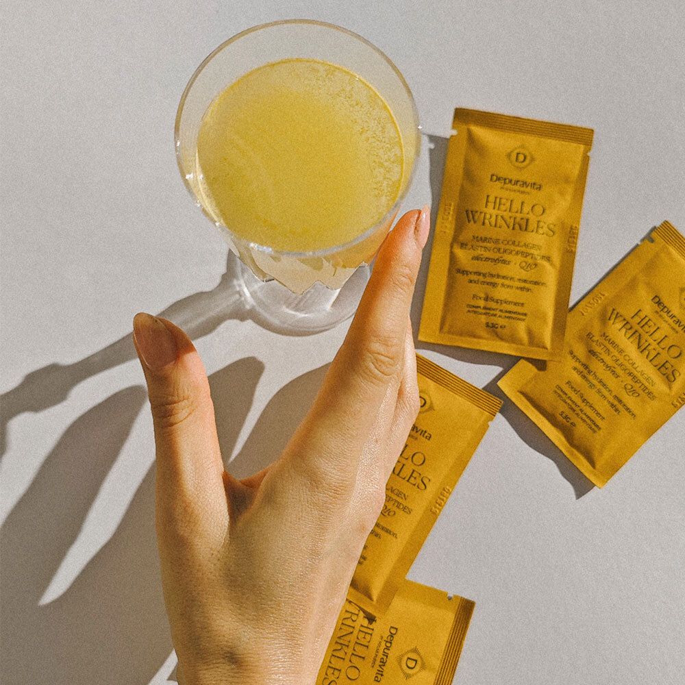 Verre de liquide jaune et trois sachets Depuravita Hello Wrinkles. Main dans l'image. Nom du produit sur les sachets.