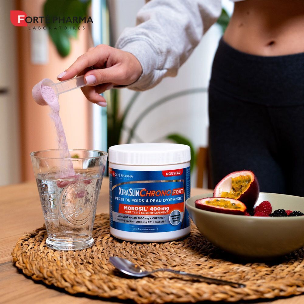 Personne verse poudre dans verre d'eau. Boîte XtraSlim Chrono Fort à côté. Fruits dans un bol. Cuillère et dessous de verre.