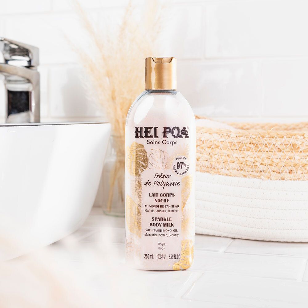 Flacon avec bouchon doré à côté d'un lavabo et d'un panier. Inscription: HEI POA, Soins Corps, Trésor de Polynésie, Lait Corps Nacré, Sparkle Body Milk.