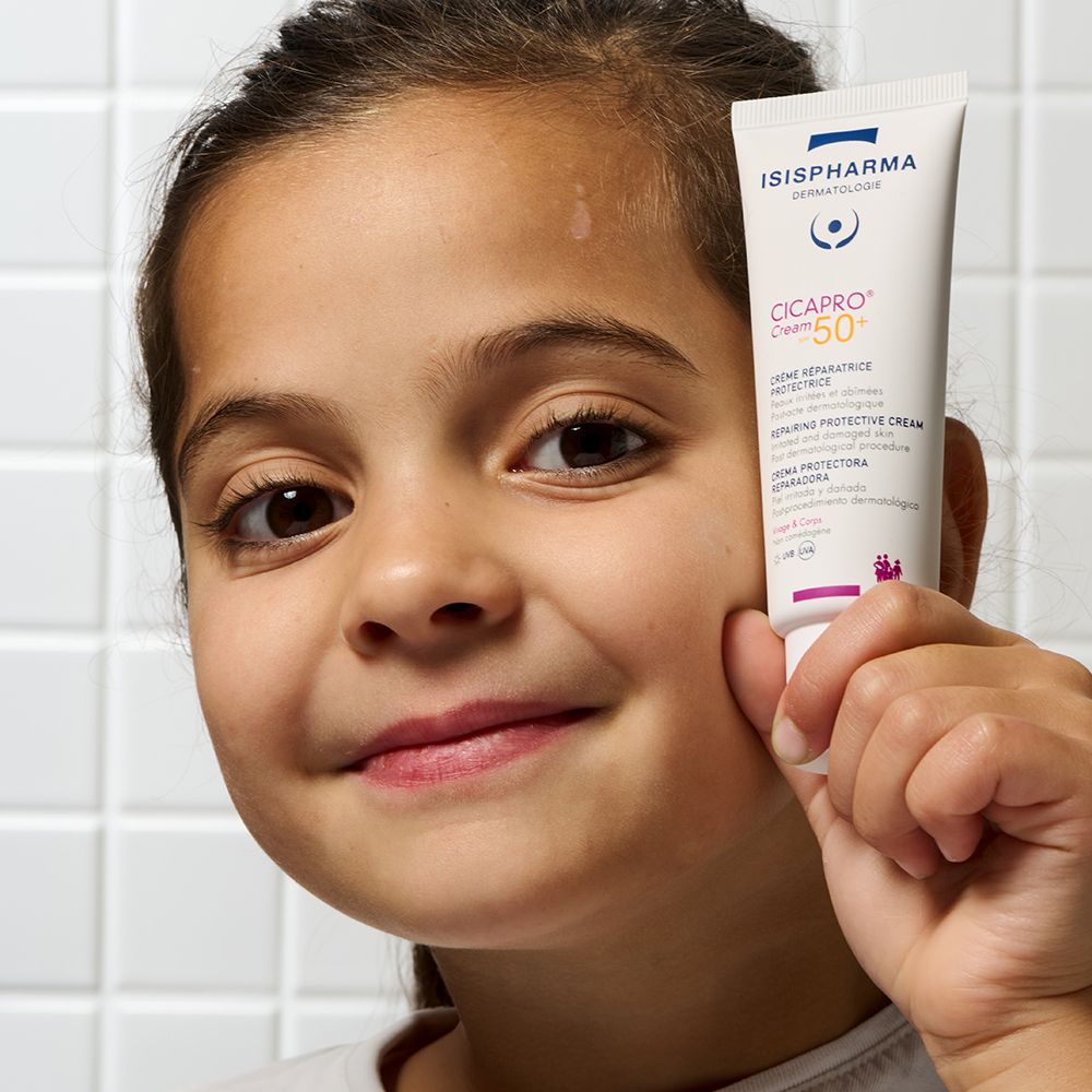Une enfant tient un tube Isispharma Cicapro Cream SPF50+ devant son visage. Gros plan.