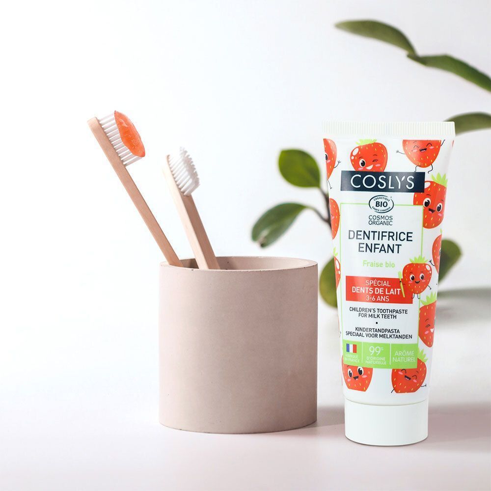 Tube de dentifrice avec motif fraise à côté de deux brosses à dents dans un gobelet. Inscription: COSLYS, DENTIFRICE ENFANT, BIO.