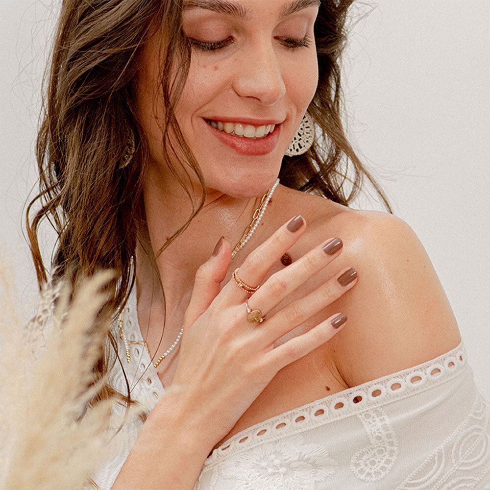 Femme avec ongles vernis marron, portant des bagues et un haut blanc. Regard vers le bas.