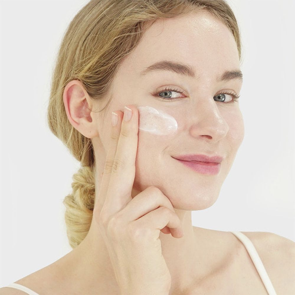 Femme appliquant de la crème sur son visage. Elle sourit. Crème sur le doigt et sur la peau. Peau claire, cheveux blonds.