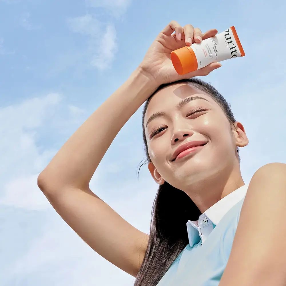 Femme tenant un tube de crème solaire au-dessus de sa tête. Ciel bleu en arrière-plan. Produit : PURITO.