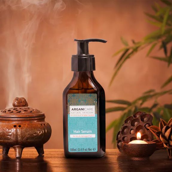 Flacon de sérum capillaire avec pompe. Inscription: Arganicare, Hair Serum. Contexte: bougie, encensoir, plantes.