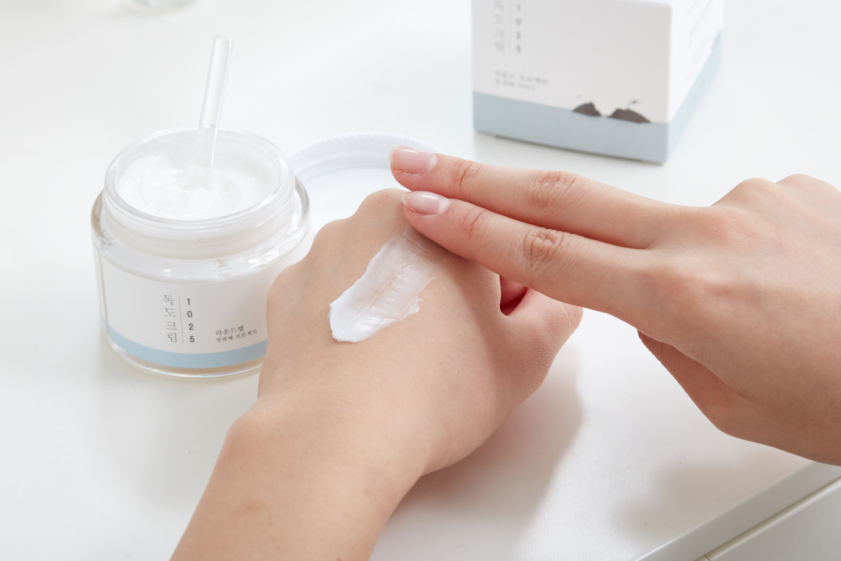 Crème pour la peau dans un pot en verre. Une main applique la crème, l'autre l'étale. Sur fond blanc.