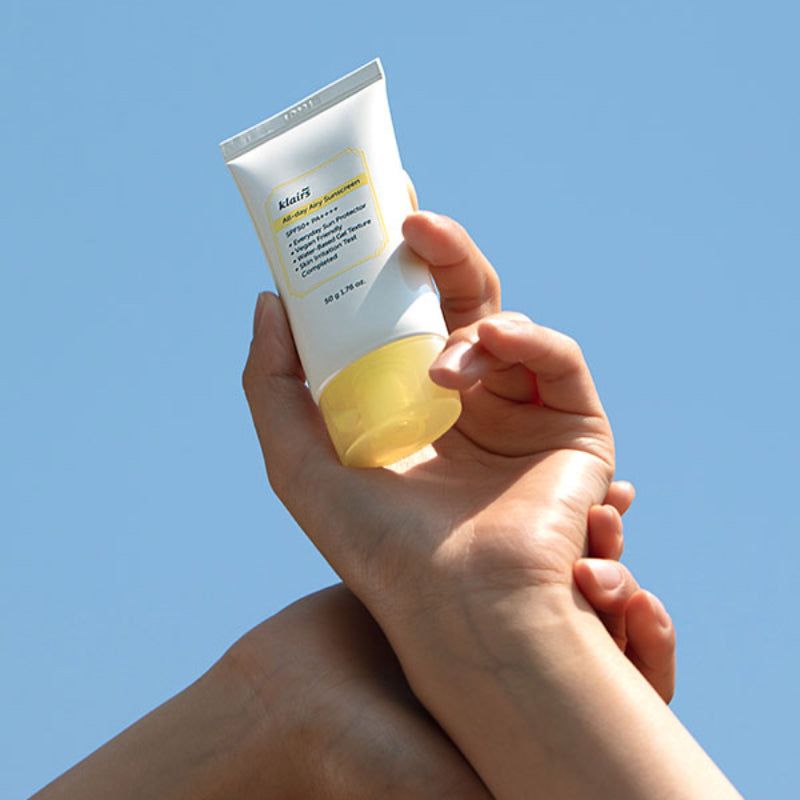 Mains tenant un tube de Klairs All-day Airy Sunscreen devant un ciel bleu.