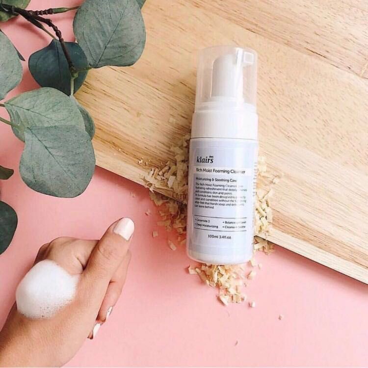Flacon Klairs Rich Moist Foaming Cleanser près d'une main avec mousse, sur bois.