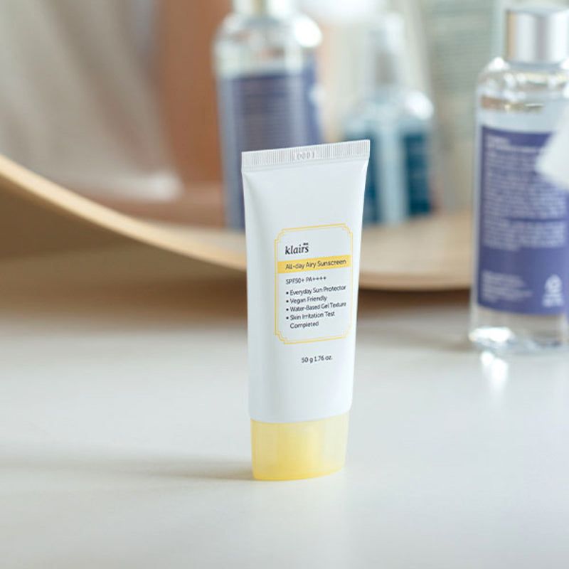 Tube Klairs All-day Airy Sunscreen sur une table, arrière-plan flou avec des bouteilles.