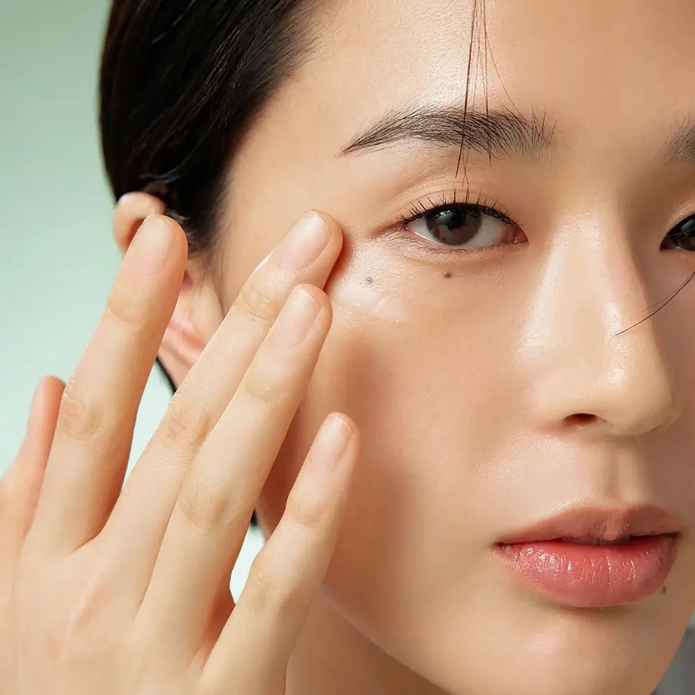 Gros plan d'une personne appliquant une crème sous l'œil. La main touche doucement la peau. Peau claire, cheveux foncés.