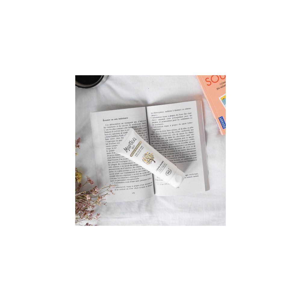 Tube blanc de crème pour les mains Marilou Bio sur livre ouvert. Logo et nom du produit visibles. Environnement: livre, fleurs.