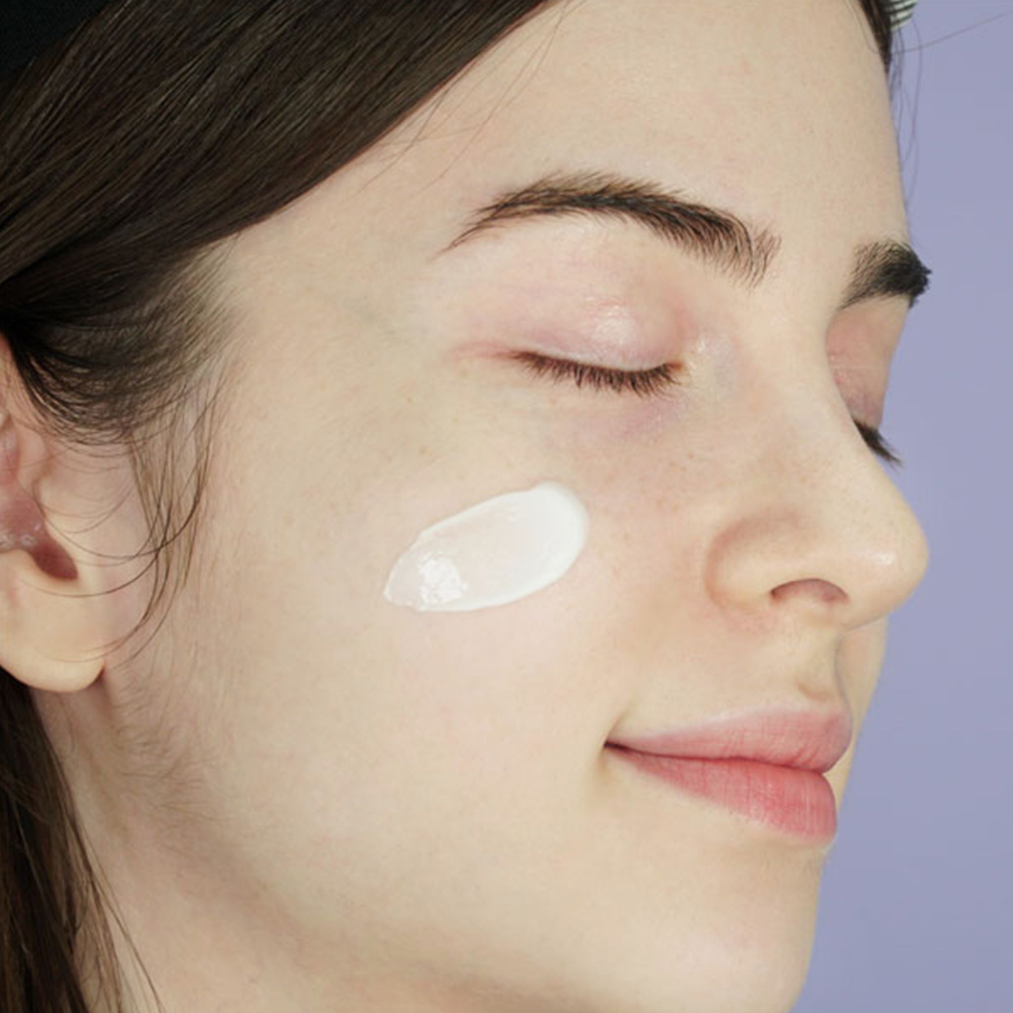 Gros plan d'une femme aux yeux fermés. Une crème blanche est appliquée sur la joue.